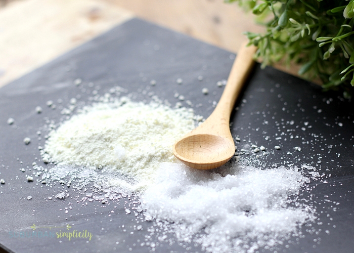 The ingredients for peppermint foot soak, salt and powdered milk, spread on a piece of gray slate. There is a wooden spoon laying next to the ingredients.