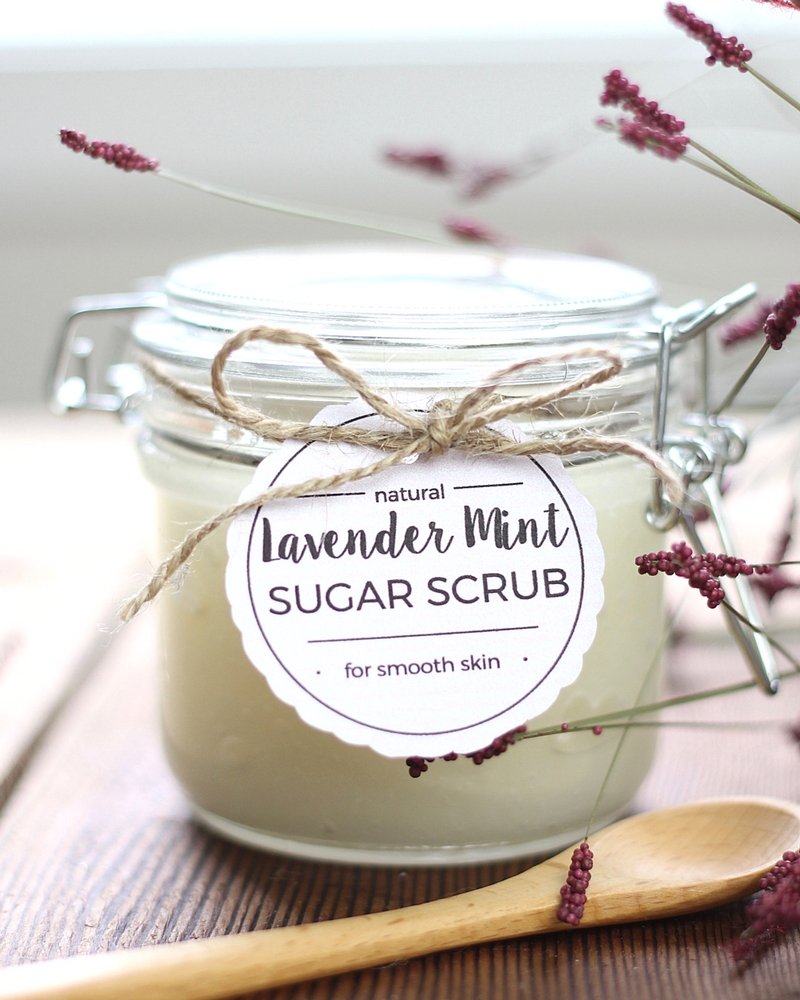 A Lavender Mint Sugar Scrub in a glass jar with a cute tag tied on with twine. A spoon sits in front and lavender flowers are in the background.