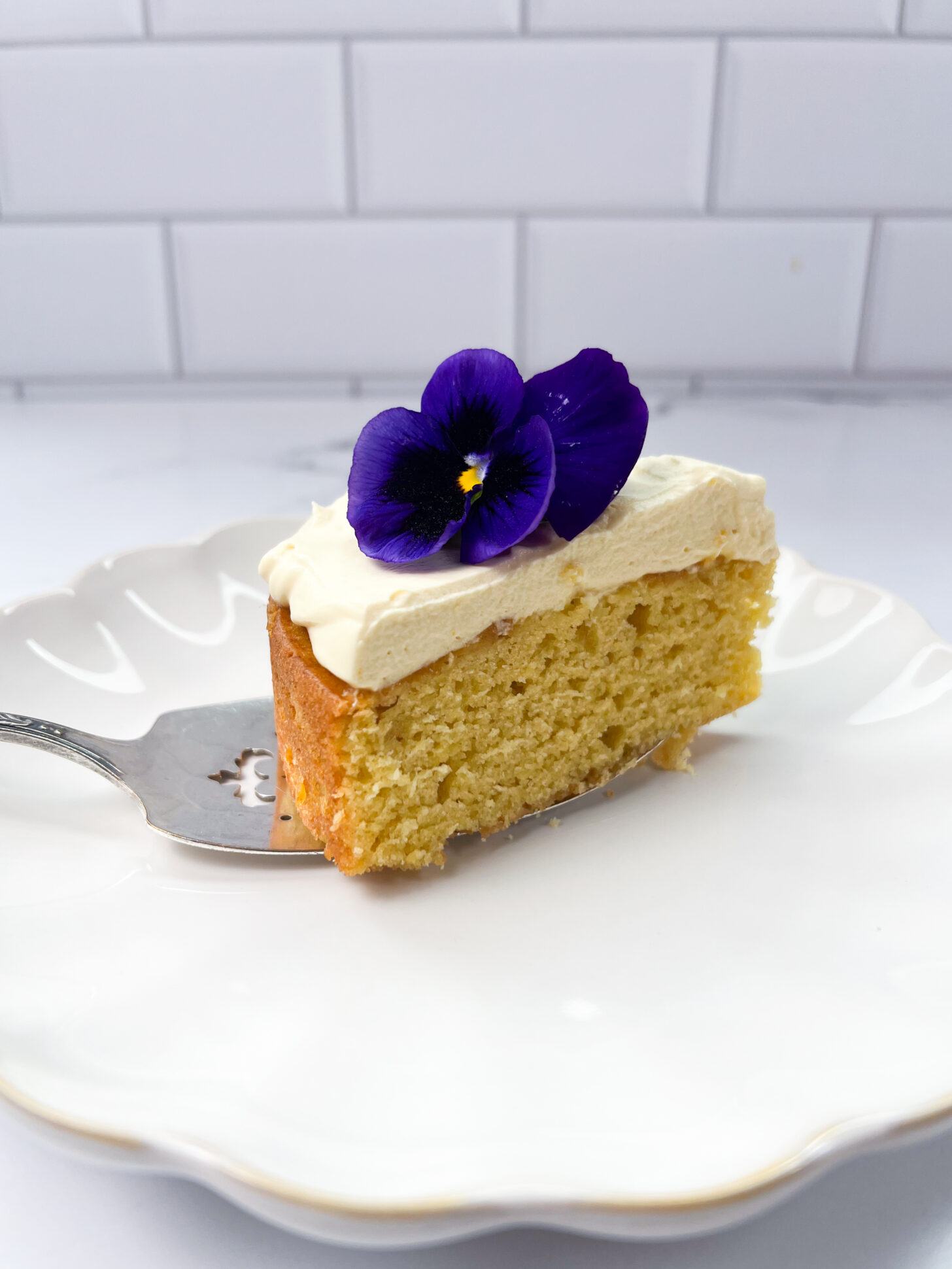 A slice of mango cake with mango mascarpone whipped cream frosting, topped with a pansy.